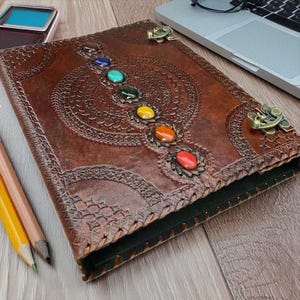 May include: A brown leather journal with a decorative embossed design and colorful gemstone accents. The journal features a series of gemstones in red, orange, yellow, green, and blue. The journal has metal corner accents.