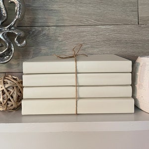 Könnte beinhalten: Ein Stapel von fünf cremefarbenen Büchern, die mit einer Schnur zusammengebunden sind. Die Bücher sind auf einem weißen Regal angeordnet.