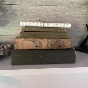 Peut inclure: Une pile de livres anciens aux tons verts, bruns et beiges. Les titres incluent "Macbeth", "Scott's Lady of the Lake" et "The Romantic Comedians". Une partition musicale roulée et attachée avec de la ficelle est posée dessus.
