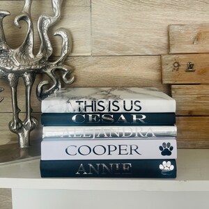 Peut inclure: Une pile de cinq livres avec des couvertures noires et blanches. Le livre du dessus a une couverture en marbre blanc avec le texte "This is Us" en noir. Le deuxième livre a une couverture noire avec le texte "Cesar" en argent. Le troisième livre a une couverture argentée avec le texte "Alejandra" en noir. Le quatrième livre a une couverture blanche avec le texte "Cooper" en argent et une empreinte de patte en argent. Le cinquième livre a une couverture noire avec le texte "Annie" en argent et une empreinte de patte en argent.