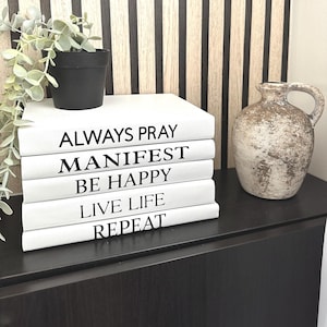 Peut inclure: Une pile de livres décoratifs blancs avec du texte noir qui dit "ALWAYS PRAY, MANIFEST, BE HAPPY, LIVE LIFE, REPEAT." Un petit pot de plante noir et un vase beige texturé sont également présents.