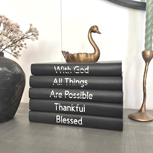 Peut inclure: Une pile de cinq livres noirs avec des lettres argentées indiquant "With God, All Things, Are Possible, Thankful, Blessed." Une figurine de cygne dorée est posée sur les livres. Un vase avec des fleurs séchées et deux bougeoirs dorés avec des bougies vertes sont également visibles.
