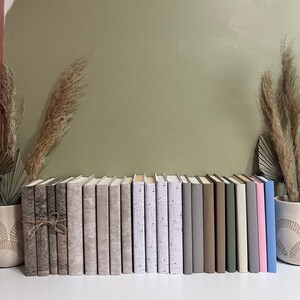 Peut inclure: Une rangée de 20 livres reliés en dur de couleur neutre avec différentes textures et motifs. Les livres sont disposés sur une surface blanche devant un mur vert.
