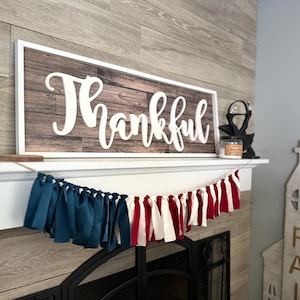May include: A wooden sign with the word "Thankful" in white script is mounted on a wall. Below, a garland of blue, white, and red fabric strips hangs above a fireplace. A decorative church-shaped item is on the right.