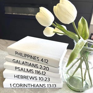Peut inclure: Une pile de livres blancs avec du texte noir, dont "PHILIPPINES 4:12" et "1 CORINTHIANS 13:13". Un vase en verre contient des tulipes blanches avec des tiges et des feuilles vertes. Les livres sont empilés sur une surface claire.