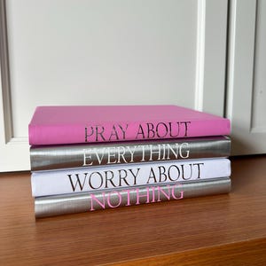 Peut inclure: Une pile de trois livres avec les phrases "PRAY ABOUT EVERYTHING" et "WORRY ABOUT NOTHING" imprimées sur les dos. Le livre du dessus est rose, celui du milieu est argenté et celui du dessous est blanc. Le texte est en rose.