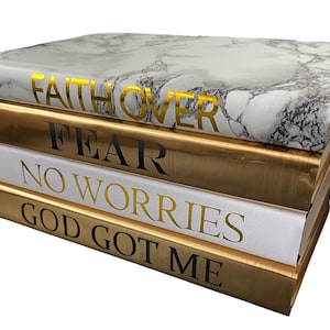 May include: A stack of five decorative books with gold and white covers. The top book has a marble pattern and the text "FAITH OVER FEAR" in gold lettering. The other books have the text "NO WORRIES" and "GOD GOT ME" in black lettering.