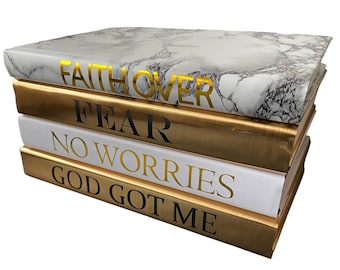 May include: A stack of five decorative books with gold and white covers. The top book has a marble pattern and the text "FAITH OVER FEAR" in gold lettering. The other books have the text "NO WORRIES" and "GOD GOT ME" in black lettering.