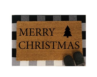 May include: A rectangular doormat made of natural coir fiber, featuring the words "MERRY CHRISTMAS" in black, along with a black Christmas tree graphic. The mat is placed on a black and white checkered rug, with a pair of gray slippers visible.