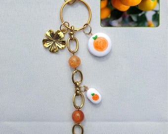 May include: A gold-colored keychain with a four-leaf clover charm, orange beads, and white charms with orange fruit designs. An inset image shows oranges on a tree. The keychain has a lobster claw clasp.
