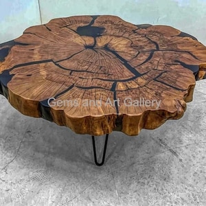Puede incluir: Una mesa de centro redonda de madera grande con un acabado de grano de madera natural y resina epoxi negra que llena las grietas. La mesa tiene una forma orgánica única y está sostenida por tres patas de horquilla negras.
