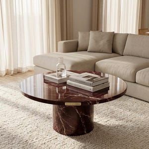 Peut inclure: Une table basse ronde en marbre bordeaux avec une base cylindrique. La table a une finition brillante et est surmont&eacute;e de livres et d'une bouteille en verre transparent. La table repose sur un tapis textur&eacute; de couleur cr&egrave;me, avec un canap&eacute; beige en arri&egrave;re-plan.