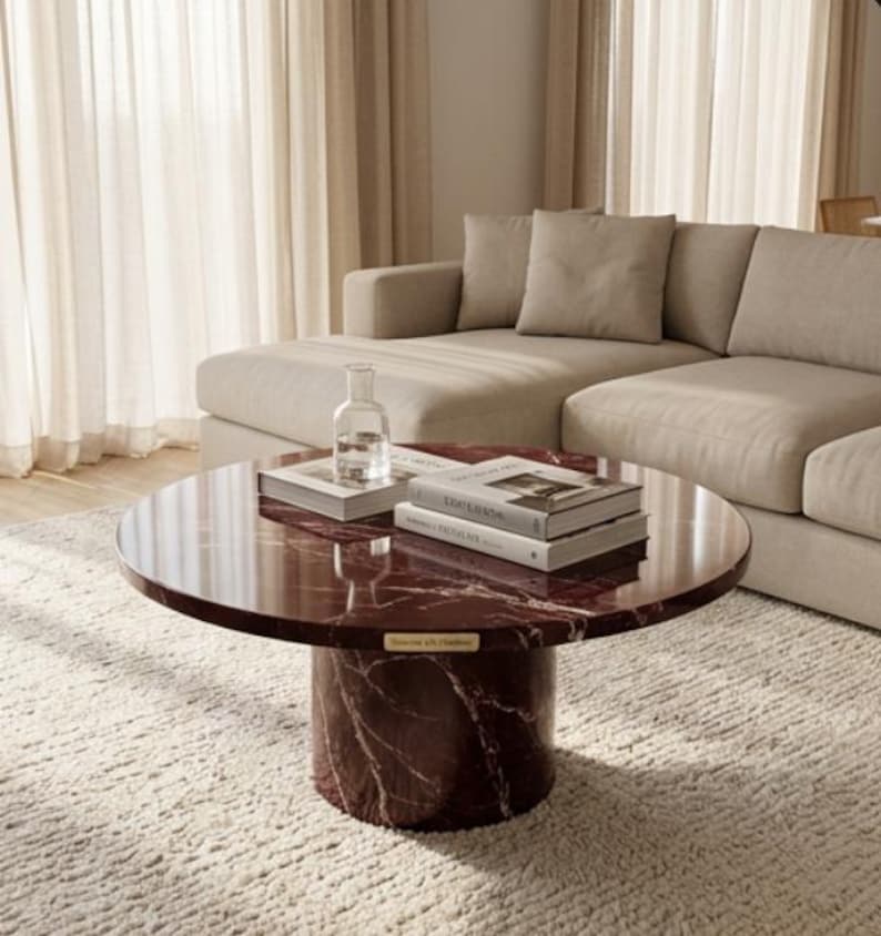 Peut inclure: Une table basse ronde en marbre bordeaux avec une base cylindrique. La table a une finition brillante et est surmont&eacute;e de livres et d'une bouteille en verre transparent. La table repose sur un tapis textur&eacute; de couleur cr&egrave;me, avec un canap&eacute; beige en arri&egrave;re-plan.