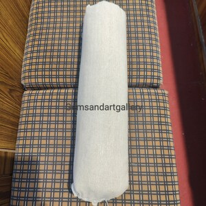Puede incluir: Una almohada cil&iacute;ndrica blanca con una superficie texturizada, descansando sobre una superficie a cuadros marr&oacute;n y negro.