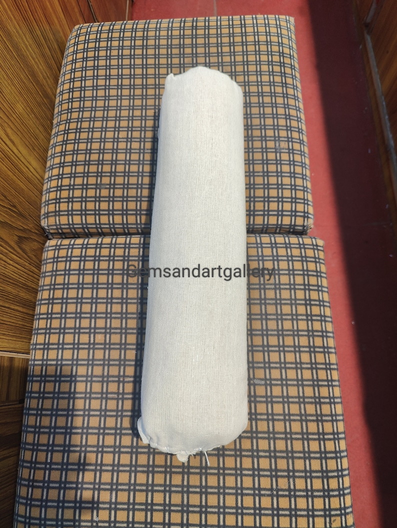 Puede incluir: Una almohada cil&iacute;ndrica blanca con una superficie texturizada, descansando sobre una superficie a cuadros marr&oacute;n y negro.