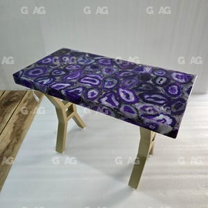 May include: A rectangular table with a polished purple agate stone top. The stone features a pattern of purple and white circular designs. The table has a gold-colored, geometric base. The table is approximately 30 inches long.