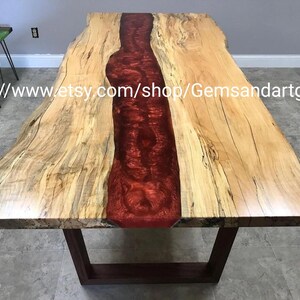 May include: A large, rectangular wooden table with a live edge design. The center of the table is filled with a vibrant red epoxy resin. The table has a dark brown base and is set on a light-colored floor.
