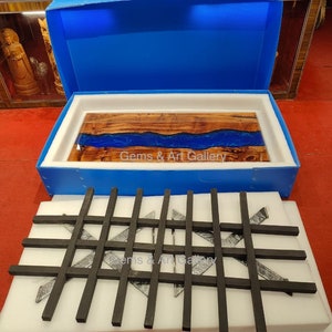 May include: A wooden table top with a blue resin river running through the center. The table top is in a blue box with white foam padding. The box is open and the table top is sitting on top of the foam. The table top is surrounded by black metal bars in a grid pattern. The text "Gems & Art Gallery" is visible on the table top.