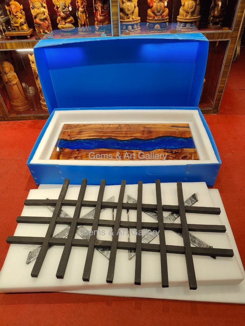 May include: A wooden table top with a blue resin river running through the center. The table top is in a blue box with white foam padding. The box is open and the table top is sitting on top of the foam. The table top is surrounded by black metal bars in a grid pattern. The text "Gems & Art Gallery" is visible on the table top.