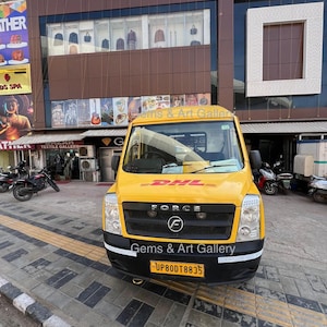 Op de afbeelding: Een gele bestelwagen met het DHL-logo en de tekst "Force" en "Gems & Art Gallery" aan de voorkant. De bestelwagen heeft een kentekenplaat met de tekst "UP80DT8835".