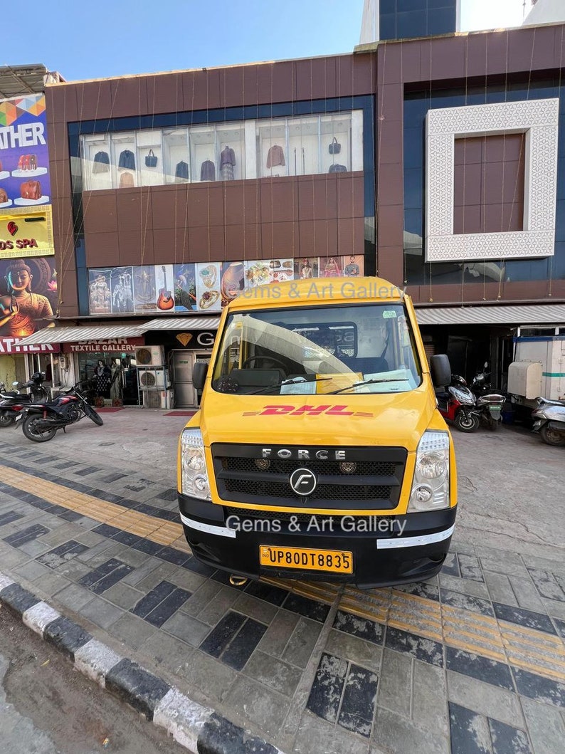 Op de afbeelding: Een gele bestelwagen met het DHL-logo en de tekst "Force" en "Gems & Art Gallery" aan de voorkant. De bestelwagen heeft een kentekenplaat met de tekst "UP80DT8835".