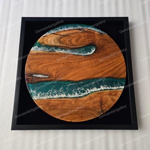 May include: A round wooden art piece with a dark frame. The wood grain is visible, with a teal and white resin design resembling waves. The artwork is a unique decorative item.