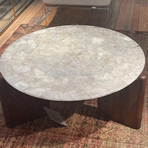 May include: Round coffee table with a white quartz top and a dark wood base. The table top is made of many small pieces of quartz that are arranged in a mosaic pattern.