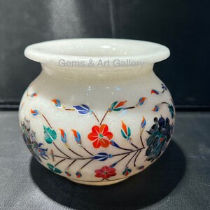 May include: A white marble vase with floral inlay. The vase features a wide mouth and a rounded body. The floral design includes red, blue, green, and orange accents. The text "Gems & Art Gallery" is visible on the vase.