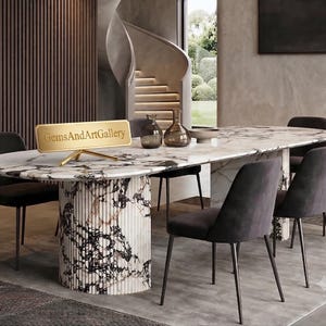 May include: An oval dining table with a white and black marble top and a fluted cylindrical base, surrounded by six dark grey upholstered chairs. A gold-toned rectangular sign with the text GemsAndArtGallery sits on the table surface.