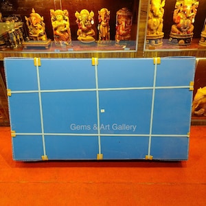以下が含まれることがあります： 白いテープと「Gems & Art Gallery」という文字が印刷された青い段ボール箱。箱は赤いカーペットの上に置かれています。