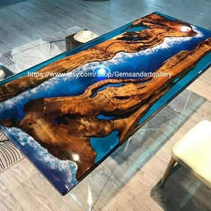 May include: A large wooden table with a blue and white epoxy resin river running through the center. The table is made of natural wood and has a unique, organic shape. The epoxy resin is clear and allows the wood grain to show through. The table is perfect for a dining room or living room.