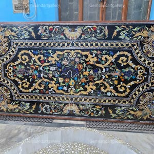 May include: A large, rectangular black marble table with intricate floral and foliate inlay in gold, blue, red, and green. The table has a dark brown border and is a statement piece for home decor.