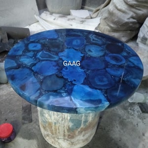 May include: A round table top featuring a mosaic of blue agate slices with varying shades of navy, cobalt, and light blue, set in a clear resin finish. The word GAAG is printed in white in the center of the circular surface.