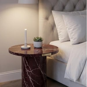 Peut inclure: Petite table de chevet ronde avec un plateau en marbre bordeaux et une base cylindrique. Un petit pot de succulente blanche et une bougie blanche sont pos&eacute;s sur la table, &agrave; c&ocirc;t&eacute; d'un lit avec une literie blanche.