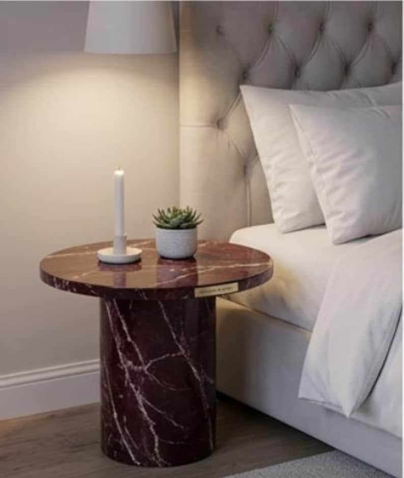 Peut inclure: Petite table de chevet ronde avec un plateau en marbre bordeaux et une base cylindrique. Un petit pot de succulente blanche et une bougie blanche sont pos&eacute;s sur la table, &agrave; c&ocirc;t&eacute; d'un lit avec une literie blanche.