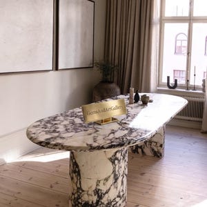 Pode incluir: Uma mesa de jantar oval feita de mármore branco com veios pretos proeminentes, apoiada por dois pedestais cilíndricos grossos de mármore. Uma placa retangular dourada com o texto GemsAndArtGallery está sobre a superfície da mesa.