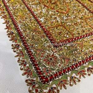 Puede incluir: Un fondo de tela blanco con un dise&ntilde;o bordado en rojo, dorado y verde. El dise&ntilde;o presenta intrincados patrones florales y peque&ntilde;as piedras preciosas de colores.