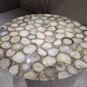 May include: A round table top constructed from numerous polished, translucent white and gray agate slices. The stones are arranged in a mosaic pattern, joined together with thin gold-colored metallic grout lines, creating a smooth, circular surface.