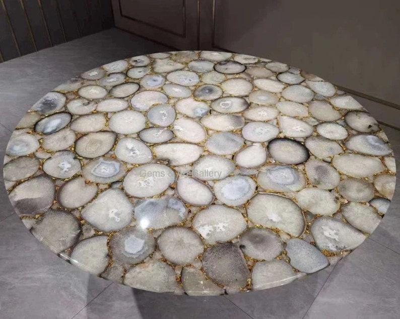 May include: A round table top constructed from numerous polished, translucent white and gray agate slices. The stones are arranged in a mosaic pattern, joined together with thin gold-colored metallic grout lines, creating a smooth, circular surface.