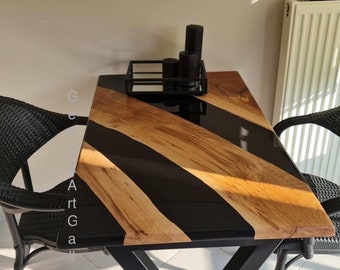 May include: A rectangular dining table featuring a natural wood grain surface with diagonal black resin inlays. The table is supported by a black metal X-frame base and is accompanied by two black woven chairs. A black metal tray holds three black candles.