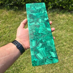 May include: A rectangular malachite stone slab with a vibrant green and black pattern. The polished surface displays a mosaic-like design, showcasing the natural beauty of the gemstone. The slab is held in a hand, with a black watch visible.