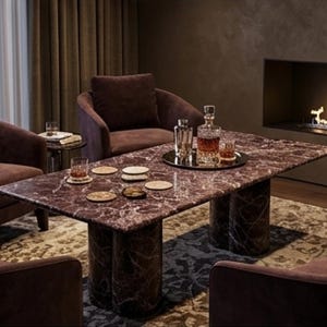 May include: A luxurious living room setting featuring a rectangular marble coffee table with cylindrical legs, surrounded by plush, dark brown armchairs. A decanter, glasses, and coasters are arranged on the table. A fireplace adds warmth to the room.