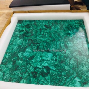 May include: A green malachite tabletop in a cardboard box with white foam padding. The tabletop has a unique, natural pattern and is ready to be shipped. The text "Gemsandartgallery" is visible in the image.