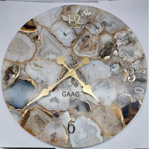 May include: A round wall clock with a white agate stone face and gold-colored hands. The clock face has Roman numerals and the word "GAAG" printed on it.
