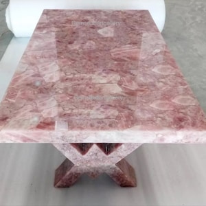 Op de afbeelding: Een roze kwarts salontafel met een X-vormig onderstel. Het tafelblad is gemaakt van gepolijst roze kwarts met een natuurlijk, onregelmatig patroon. Het onderstel is ook gemaakt van roze kwarts en heeft een ruwe, onafgewerkte uitstraling.