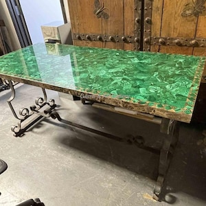 Puede incluir: Una mesa con tapa de malaquita verde con un borde decorativo, sostenida por una base de hierro forjado. La mesa está en una habitación con una puerta de madera en el fondo.