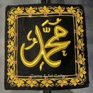 Puede incluir: Un cuadrado de terciopelo negro con un diseño de caligrafía árabe bordado en oro. La caligrafía está rodeada por un borde floral dorado y verde con joyas rojas.