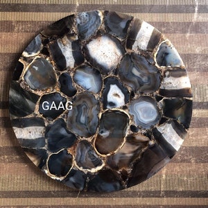 Puede incluir: Una bandeja o posavasos decorativo circular hecho de rodajas de piedra ágata pulida en negro, gris y blanco dispuestas en un patrón de mosaico con bordes dorados. La palabra GAAG está impresa en letras blancas en el lado izquierdo de la superficie.
