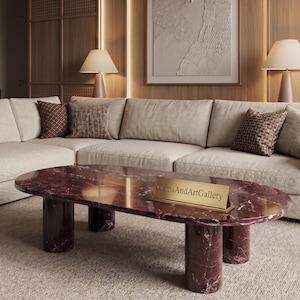 Peut inclure: Une table basse ovale en marbre bordeaux profond avec une surface polie et quatre pieds cylindriques. Une petite plaque signalétique rectangulaire dorée portant le texte GemsAndArtGallery est posée au centre de la table.