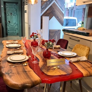 May include: A live edge wood dining table with a central translucent red resin river design, set with white plates, gold cutlery, beige napkins, and red floral centerpieces. A gold sign on the table displays the text Gemsandartgallery.